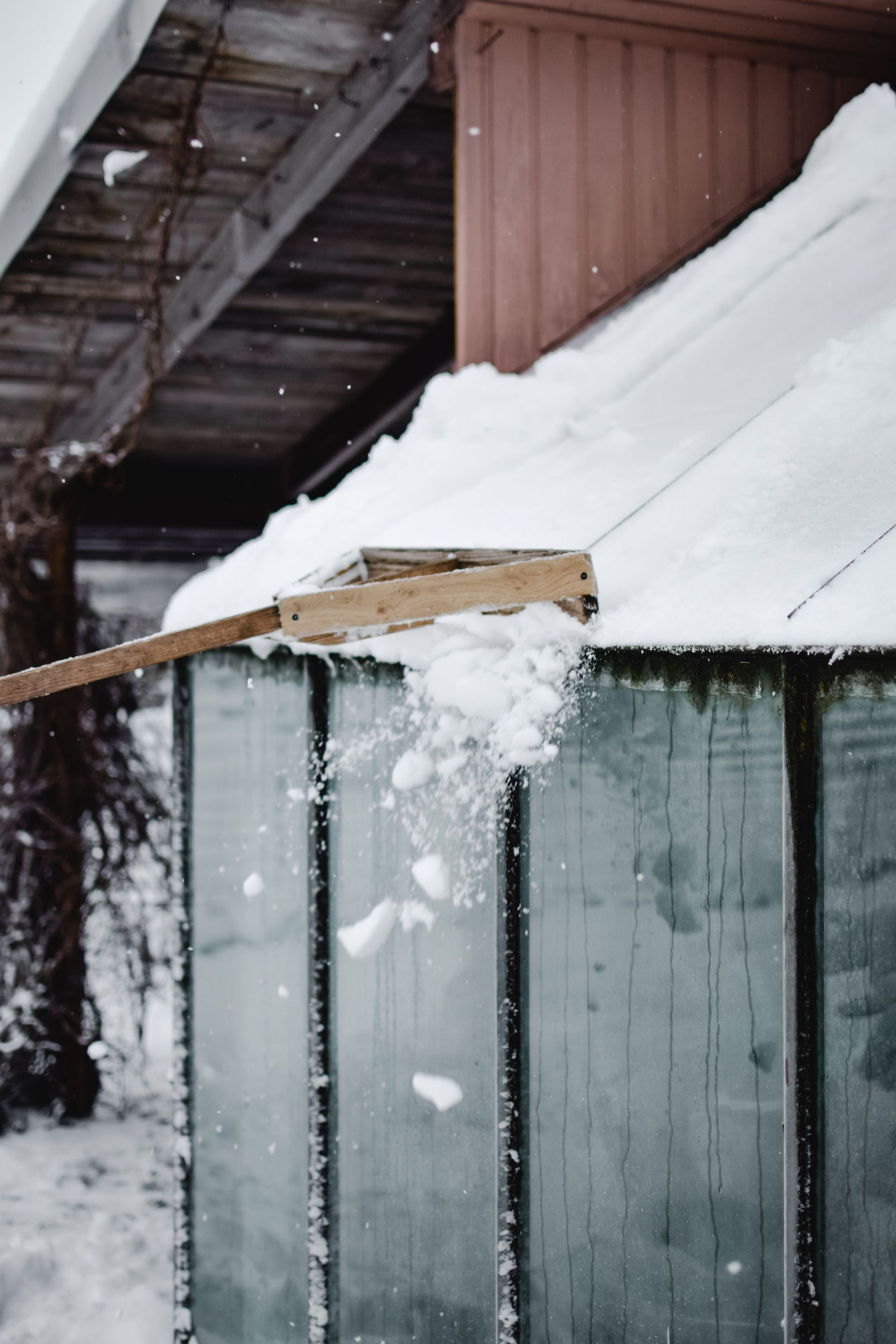 Snow piled on a roof and wooden fence beside a building, with snow falling in front.