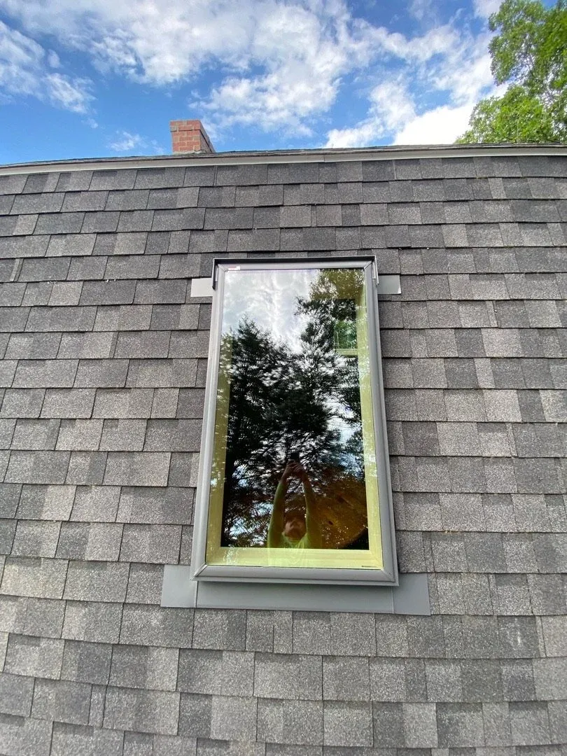 Dormer window on a gray shingled roof reflecting trees and blue sky
