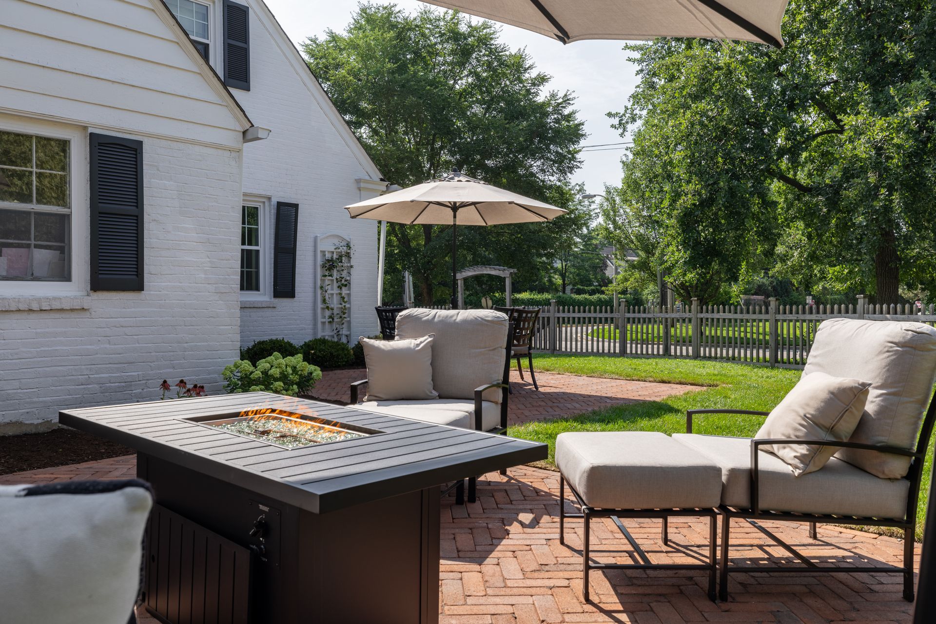 A patio with a fire pit , chairs and umbrellas in front of a white house.