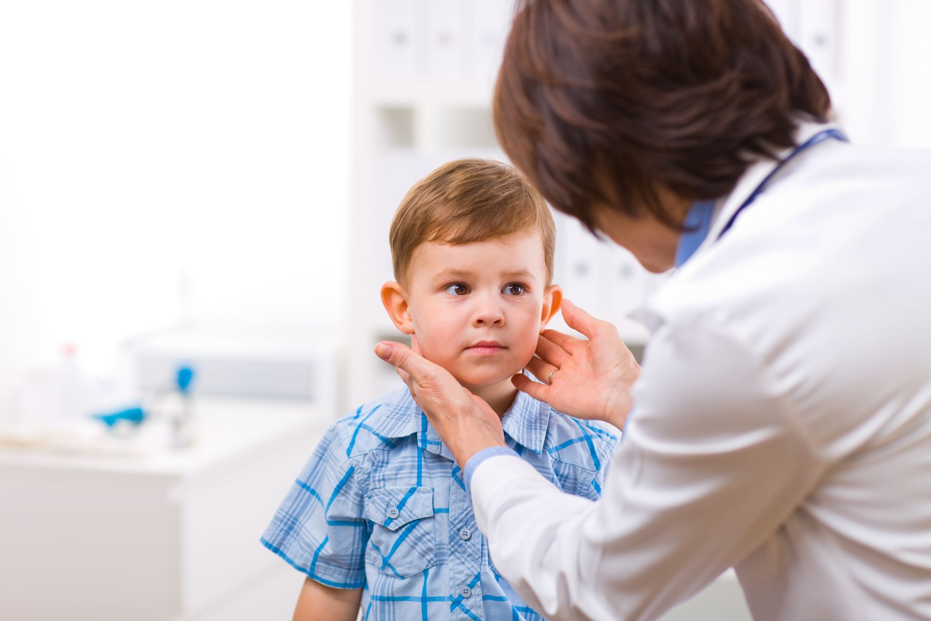A healthcare professional in a white coat examines a young child's neck in a bright clinic setting.