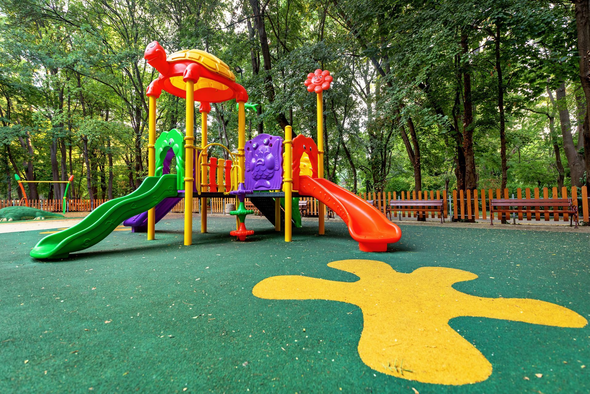 A colorful playground with a turtle-shaped roof, two slides, and a yellow splash pattern on the green ground in a park.