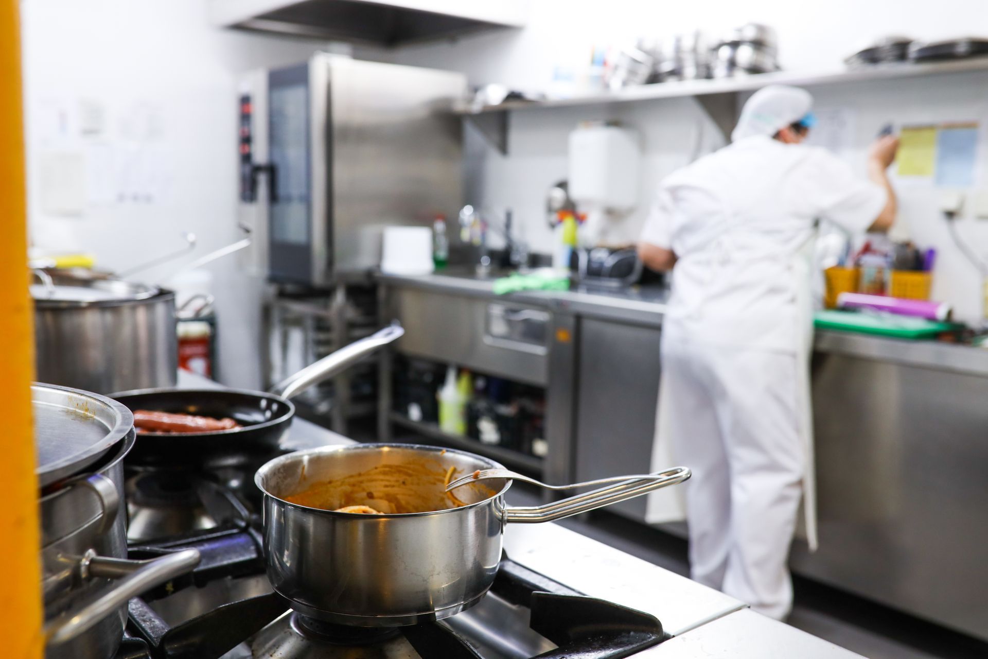 A professional kitchen with a chef in white uniform working at a counter, with a stainless steel pot on a gas stove.
