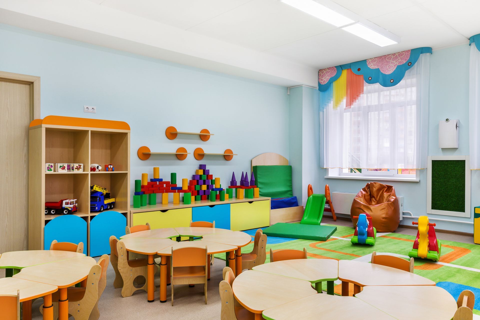 A bright, colorful preschool classroom featuring round wooden tables, child-sized chairs, shelves with toys, and a play mat.