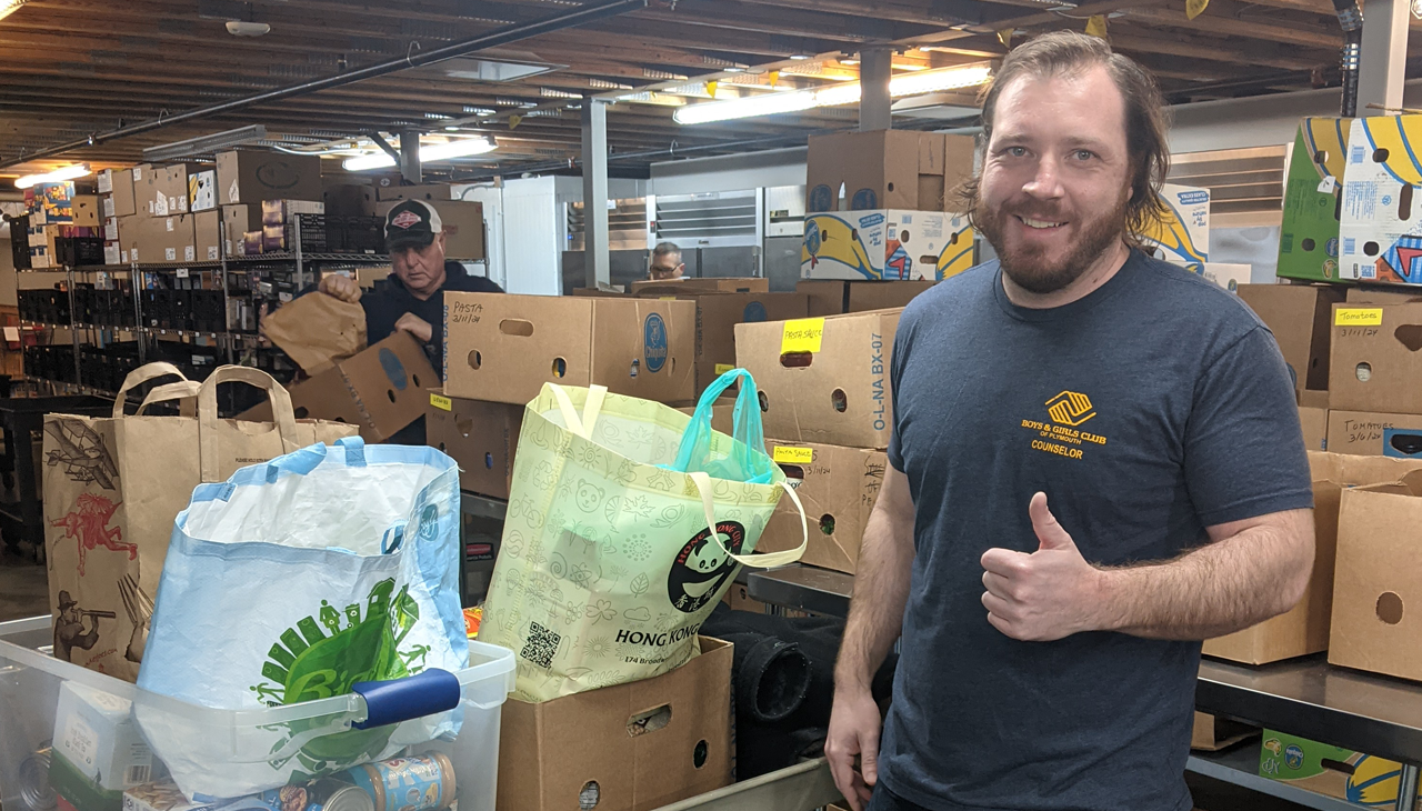 A smiling person giving a thumbs up in a warehouse filled with cardboard boxes and bags of food.