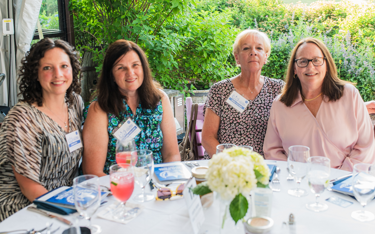 Four women seated at a table with drinks and flowers in a bright garden setting
