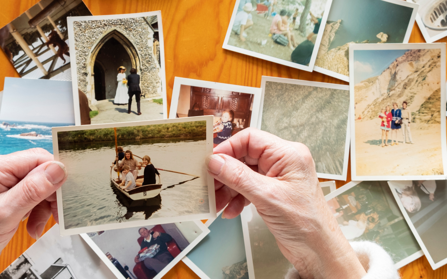 Hands hold a photo of people in a boat, surrounded by other scattered vintage photographs on a wooden table.