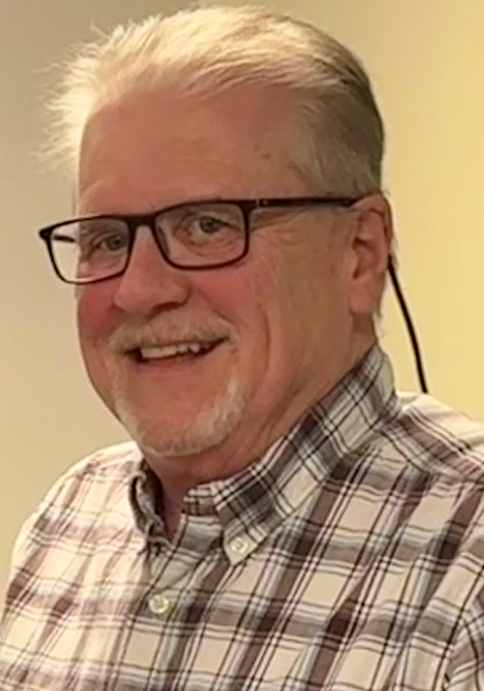Smiling man with glasses and a plaid shirt against a plain beige background