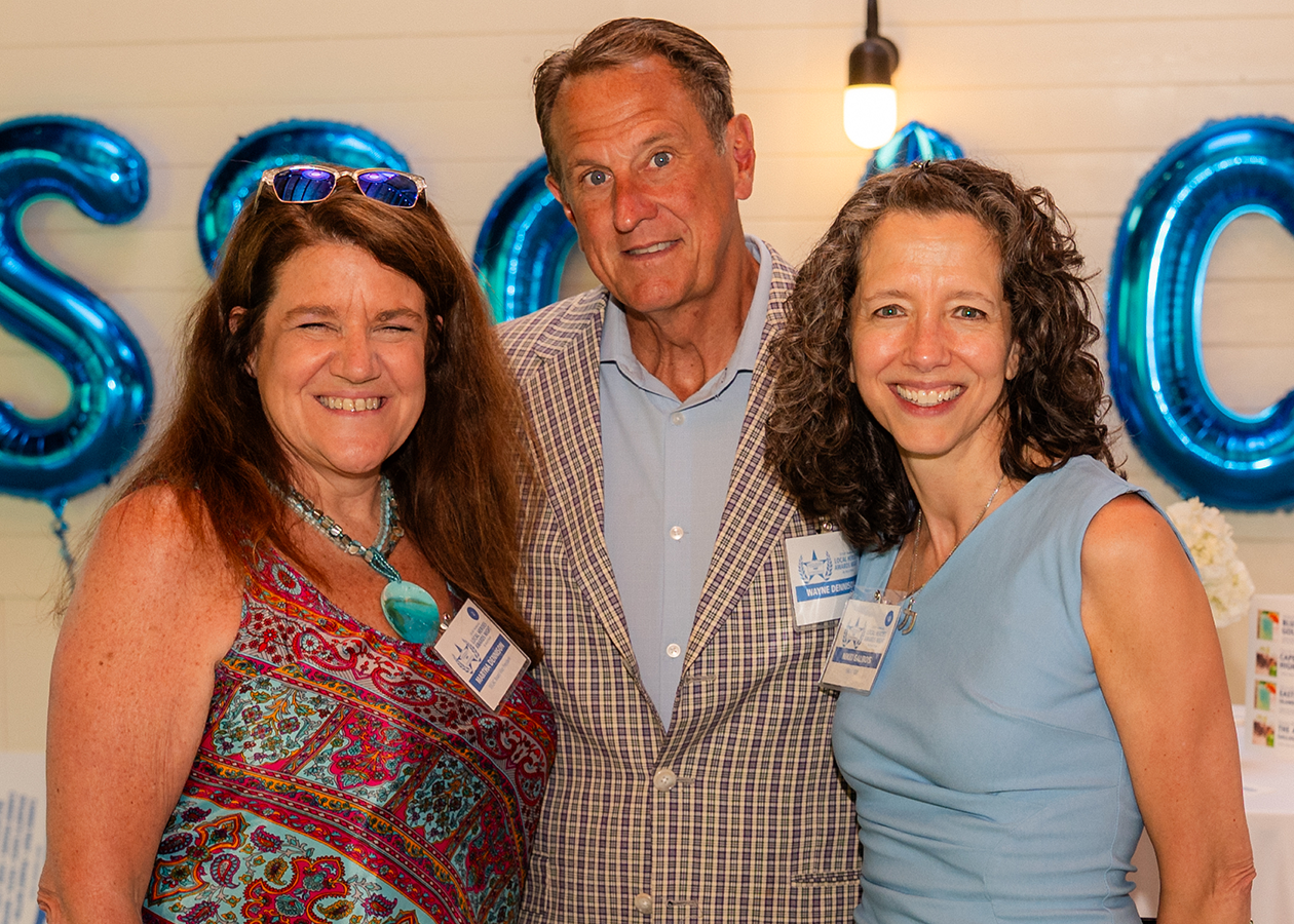 Three smiling people posing indoors at a party in front of glowing blue balloons