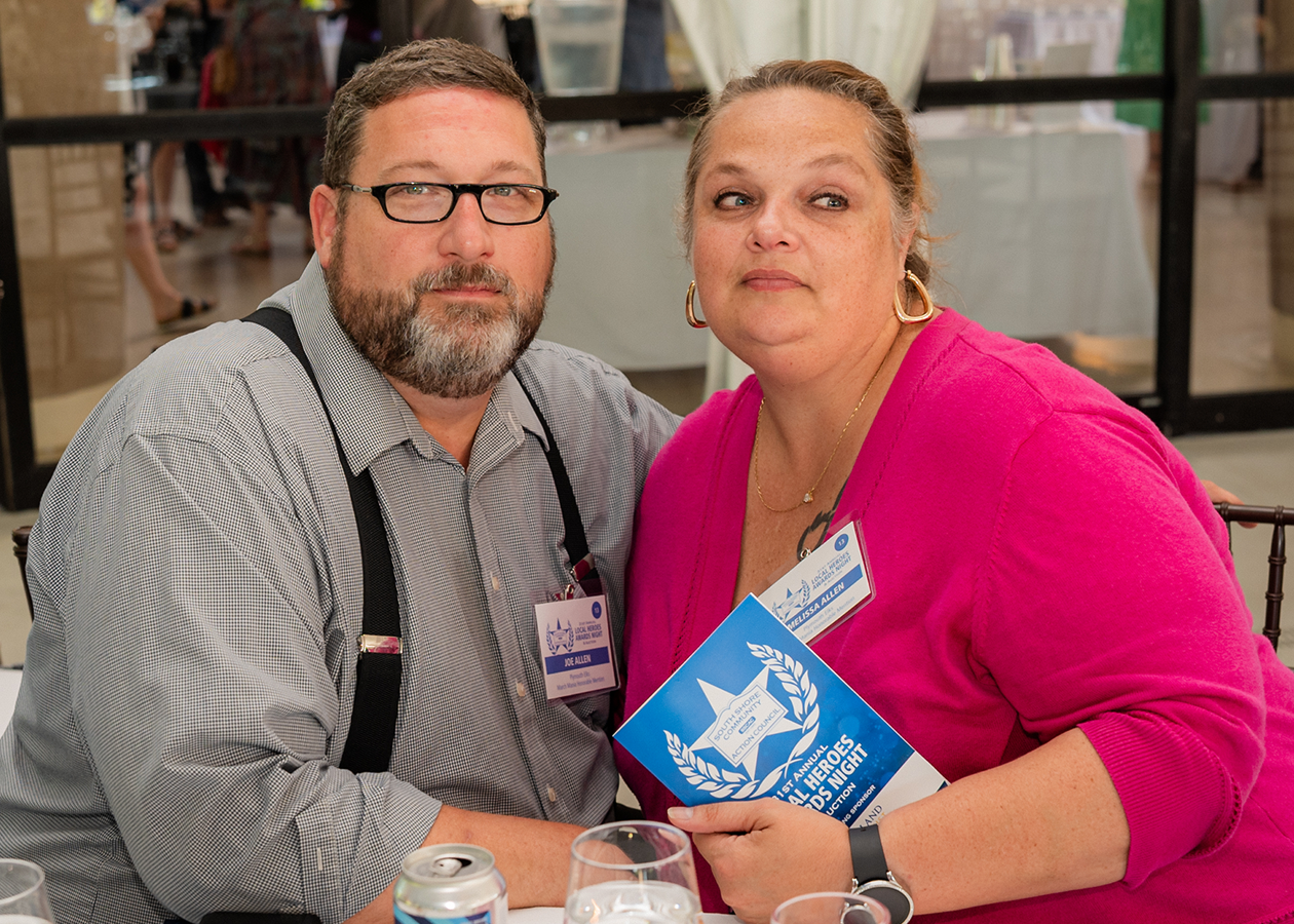 Two people seated at a table, holding a blue booklet, with name badges in an indoor event setting