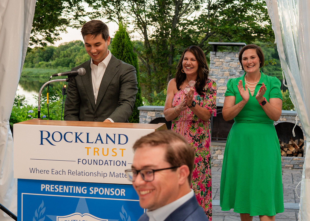 Three people on a stage, one at a Rockland Trust podium, applauding at an outdoor event.