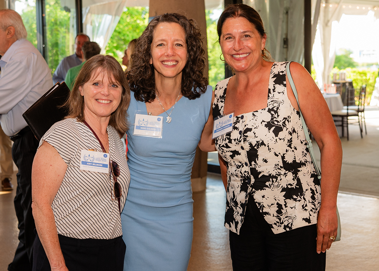 Three women smiling indoors at a social event, standing close together in front of large windows.