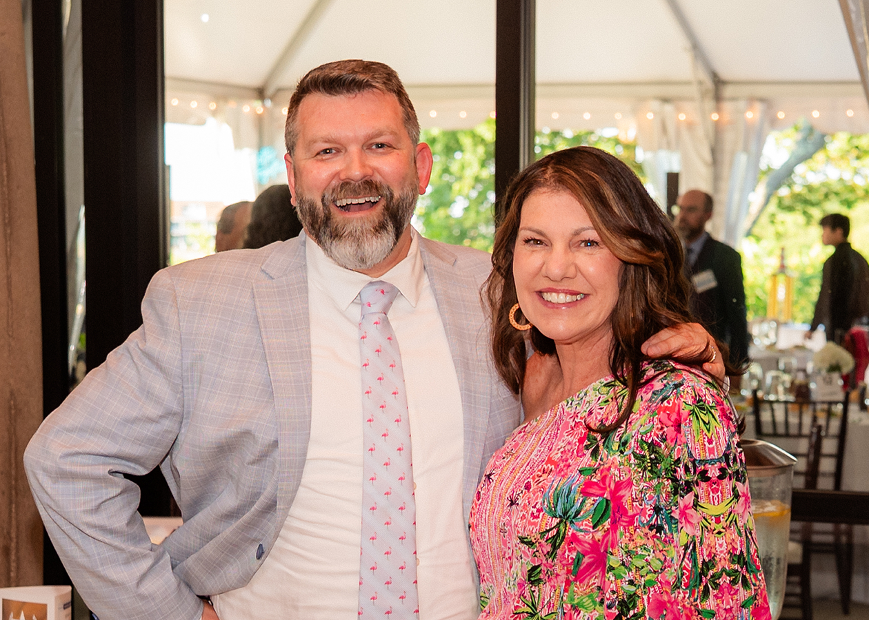 Two smiling people stand together at a tented outdoor event, wearing light-colored formal attire.