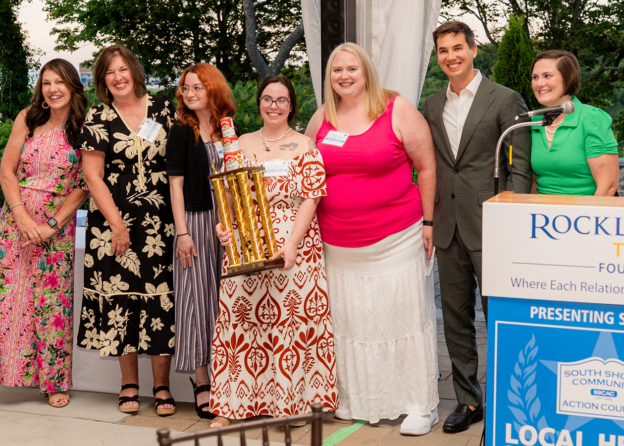 Seven people stand smiling at an outdoor awards event beside a podium and banner.