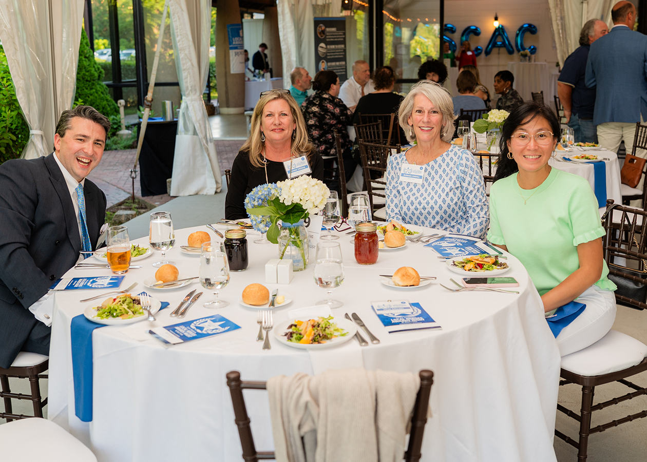 Four people seated at a round banquet table outdoors, with food, drinks, and white table linens.