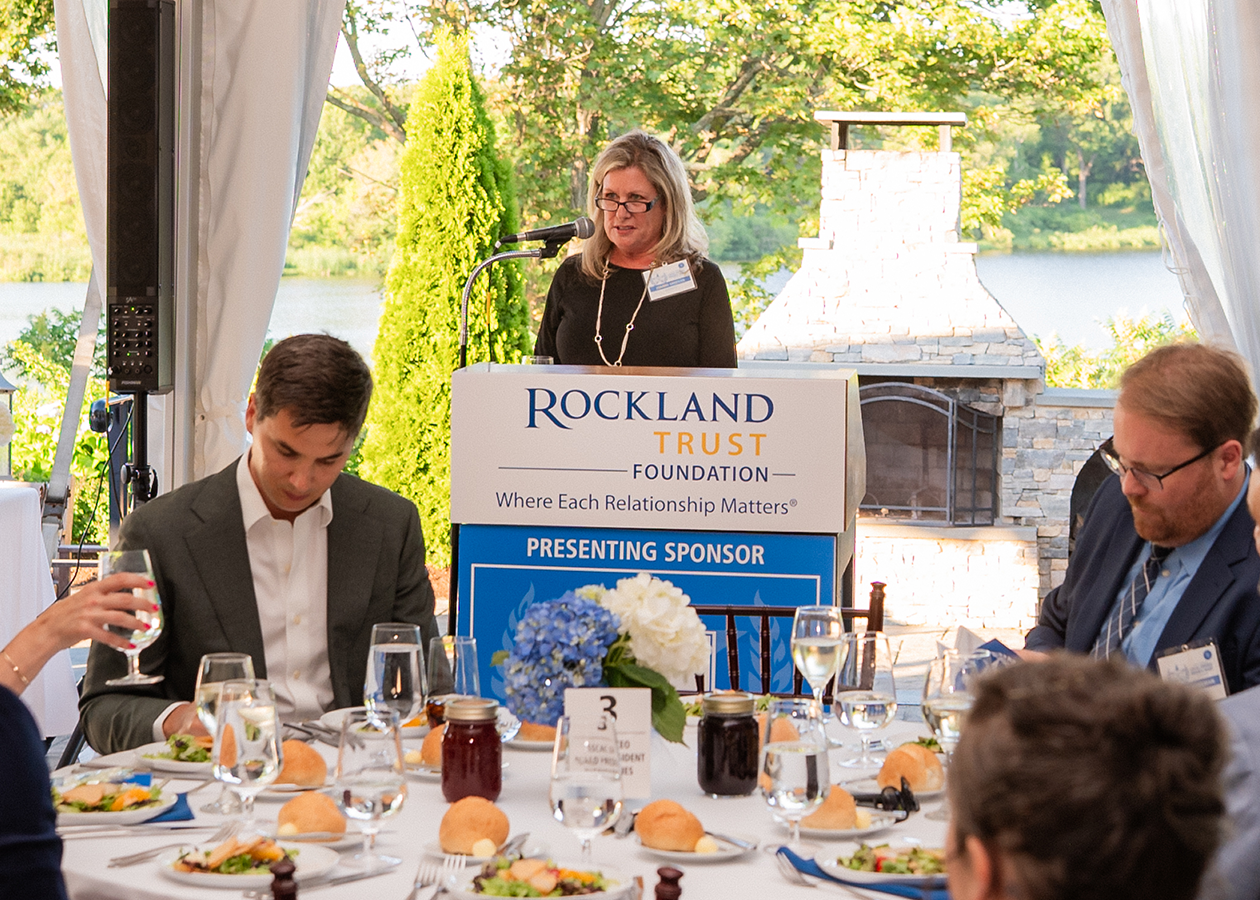 Speaker at an event addresses seated guests at a table outdoors by the water.