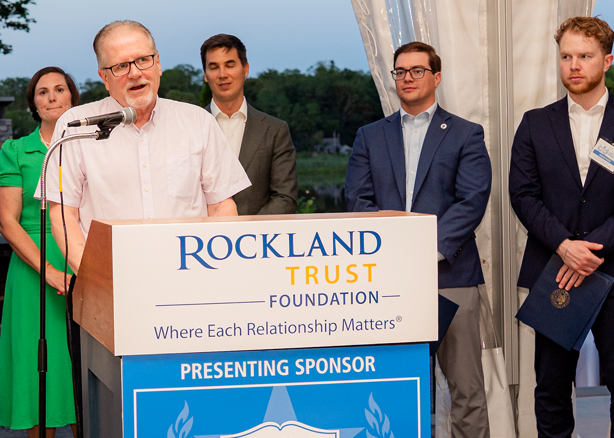 Man speaking at podium with others standing behind him outdoors at dusk