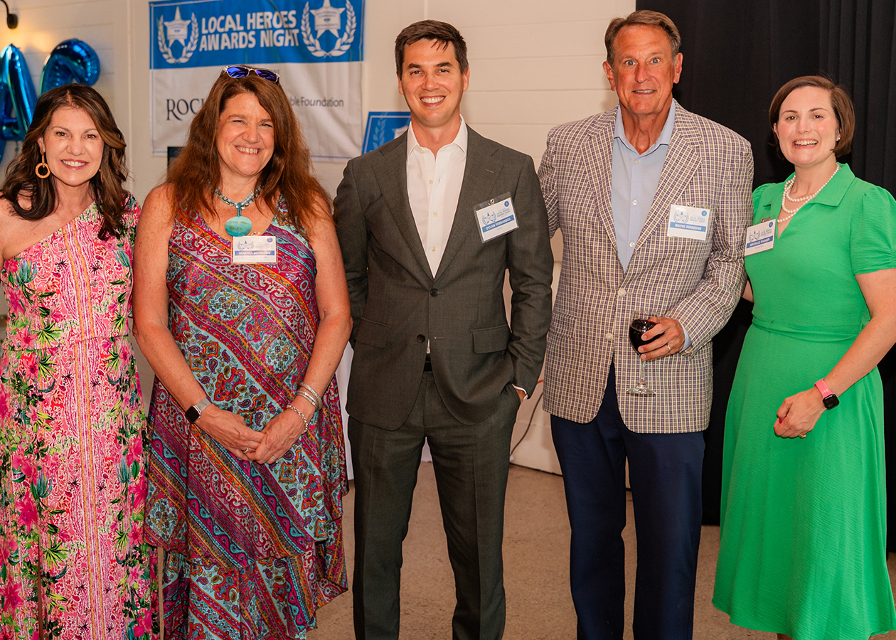 Five people posing indoors at a formal event, with a blue banner and tan backdrop behind them.