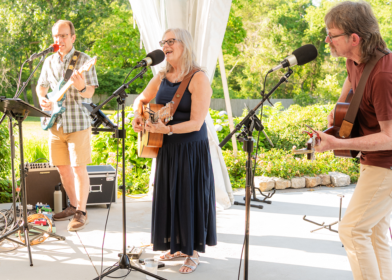 Three musicians performing outdoors on a patio, with microphones and guitars in front of green trees.