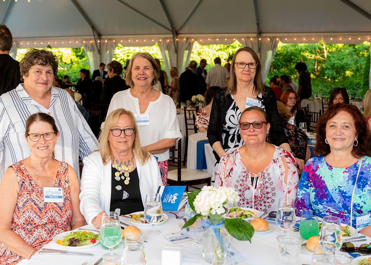 Group of women seated at a banquet table under a tent, smiling at the camera.
