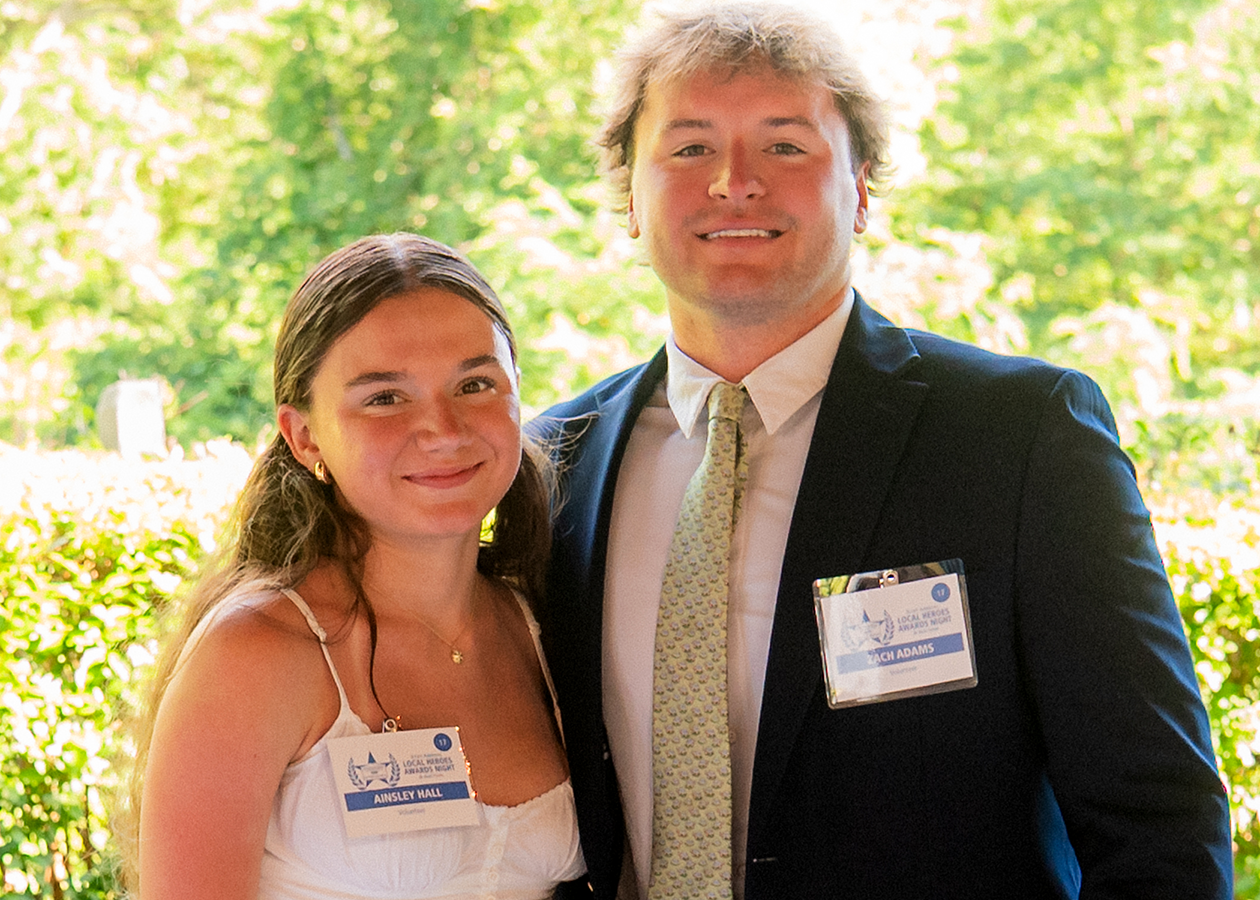 Two people smiling outdoors in formal attire with name badges, standing in a sunny garden area.
