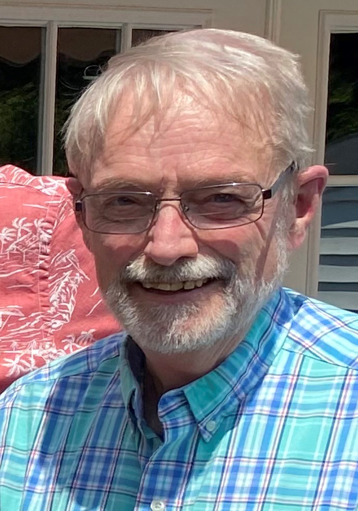 Smiling man with glasses and white hair in a blue plaid shirt, standing outdoors in sunlight