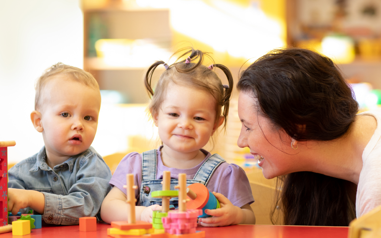 A teacher smiles as two children play with colorful stacking toys on a red table in a classroom.