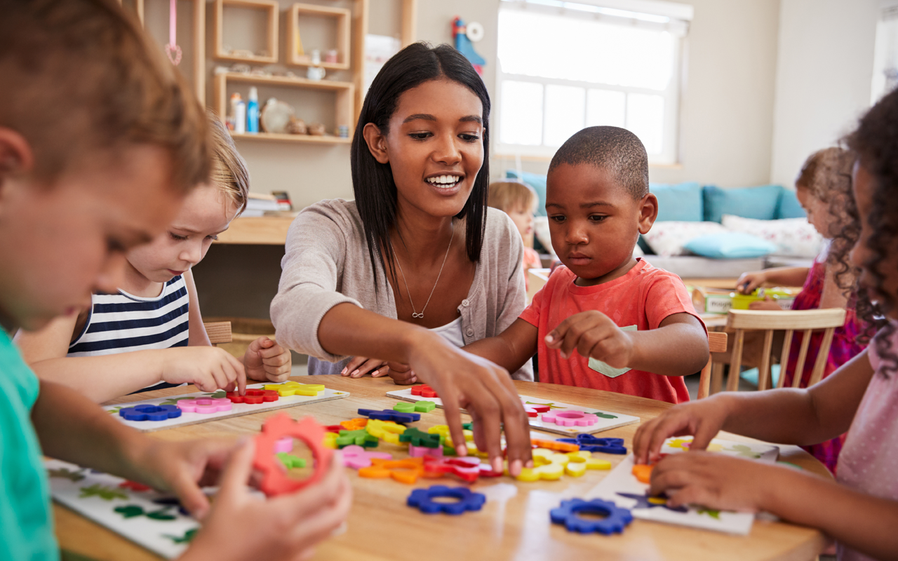 A teacher and children sit around a table playing with colorful plastic shapes in a classroom.