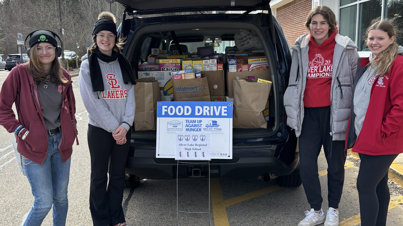 Four people stand in an outdoor parking lot behind a vehicle filled with boxes and bags for a labeled food drive.