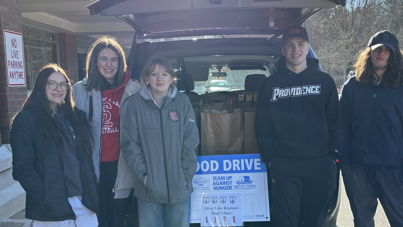 Five students stand before a car trunk filled with brown paper bags, featuring a blue sign that reads