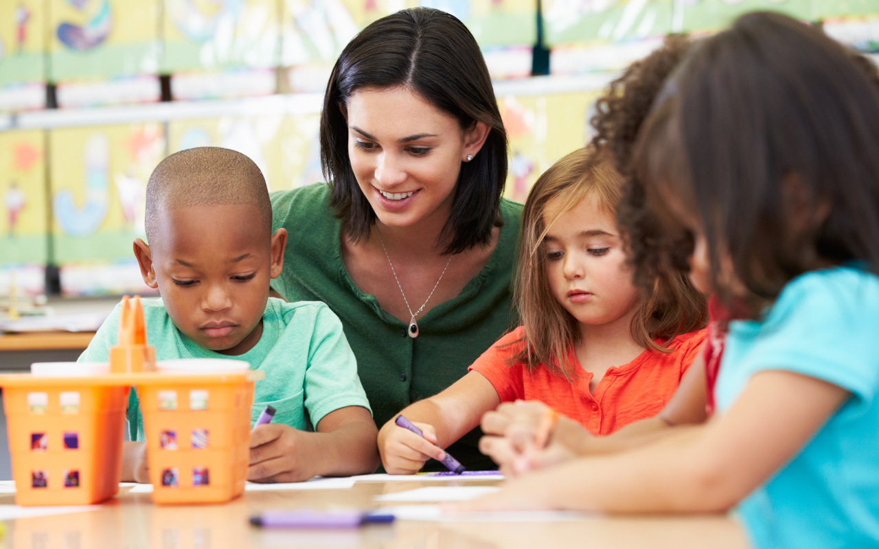A teacher smiles while sitting with three students at a table, all of whom are focused on coloring with crayons.