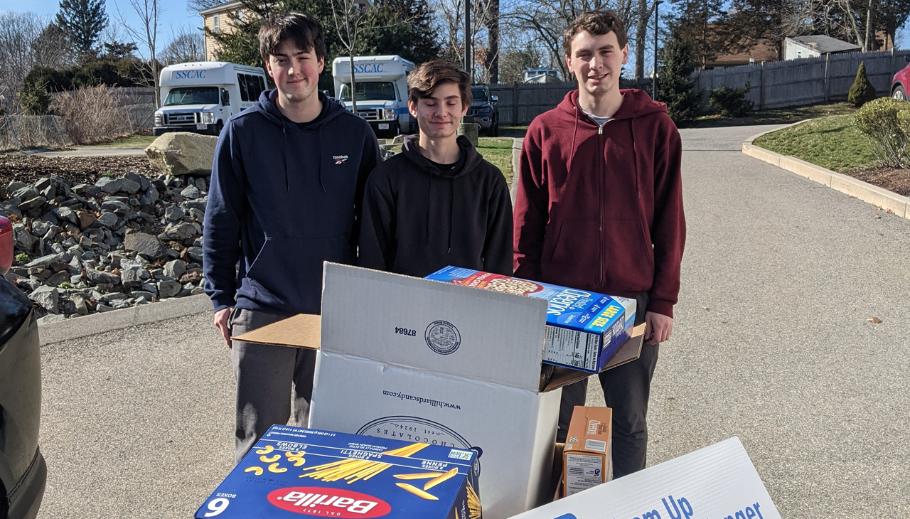 Three young people stand outdoors behind boxes of donated food, smiling near a parked recreational vehicle.