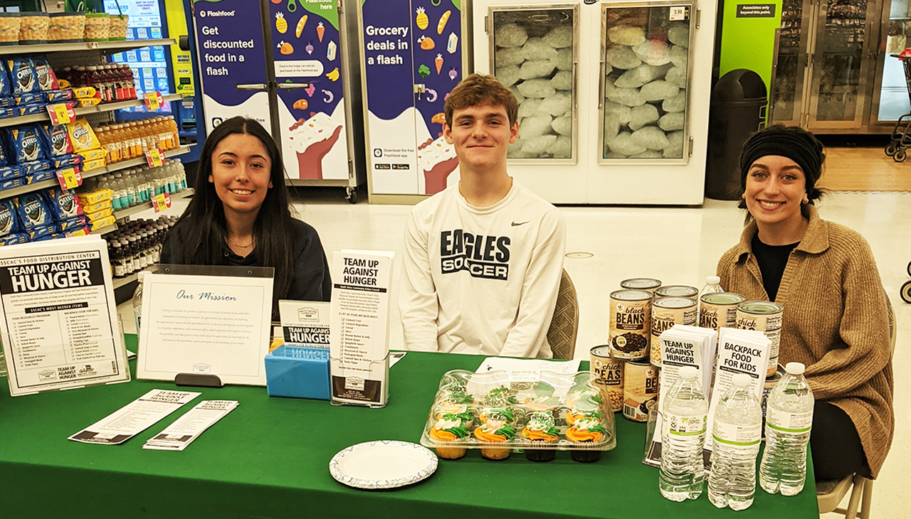 Three people sit behind a table covered in a green cloth with food, water, and signs inside a store.