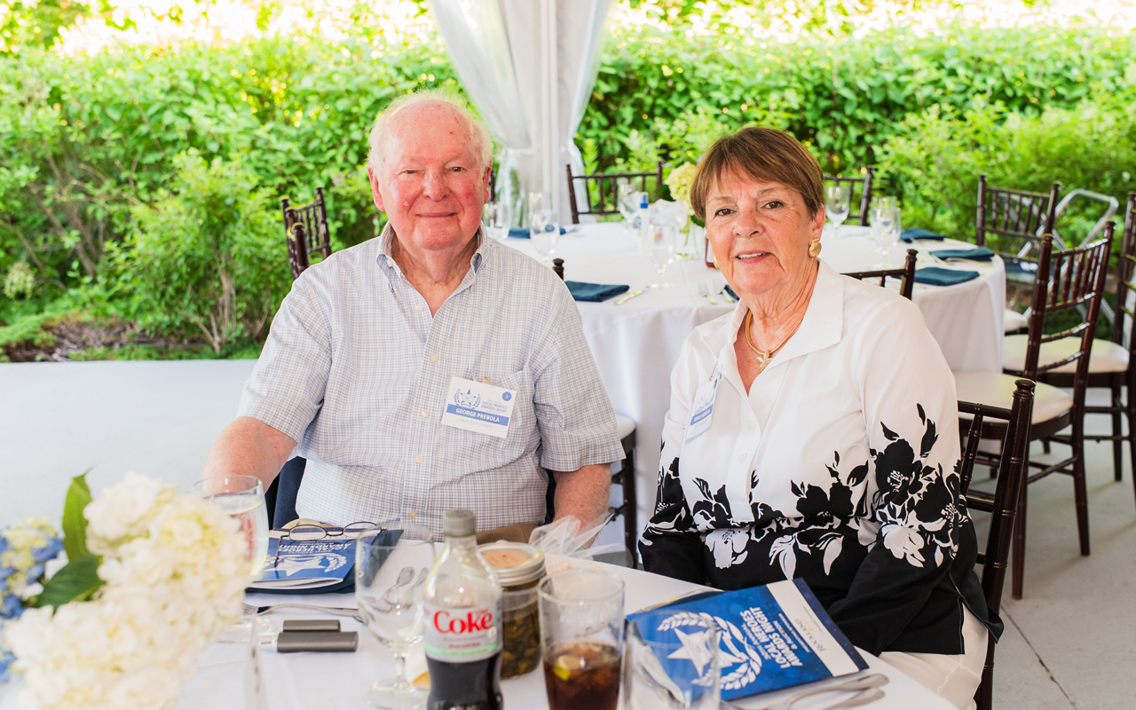 Two people seated at a table outdoors under a white canopy, smiling with drinks and books nearby.