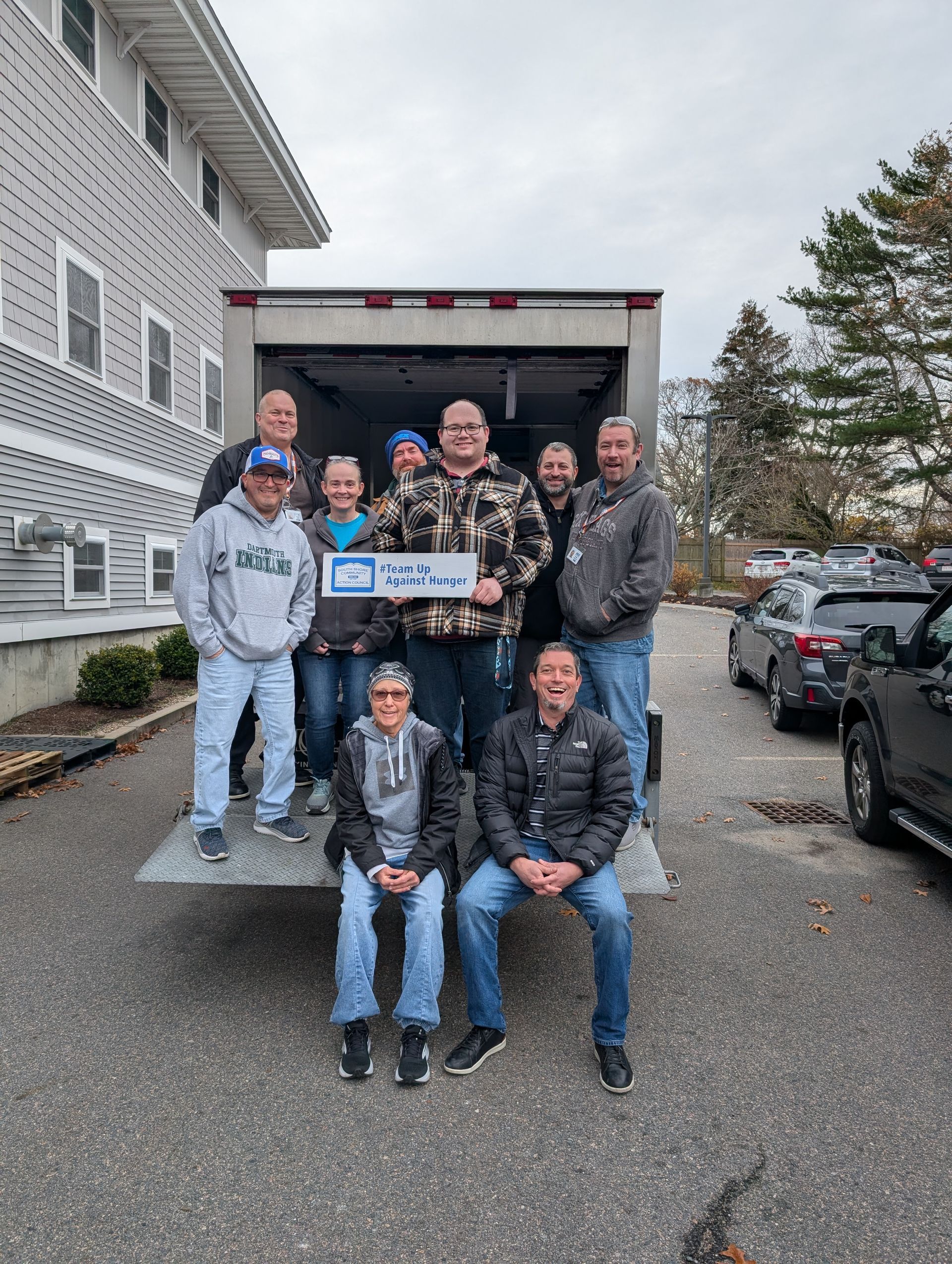 A group of people standing and sitting on the open back of a moving truck outdoors on an overcast day.