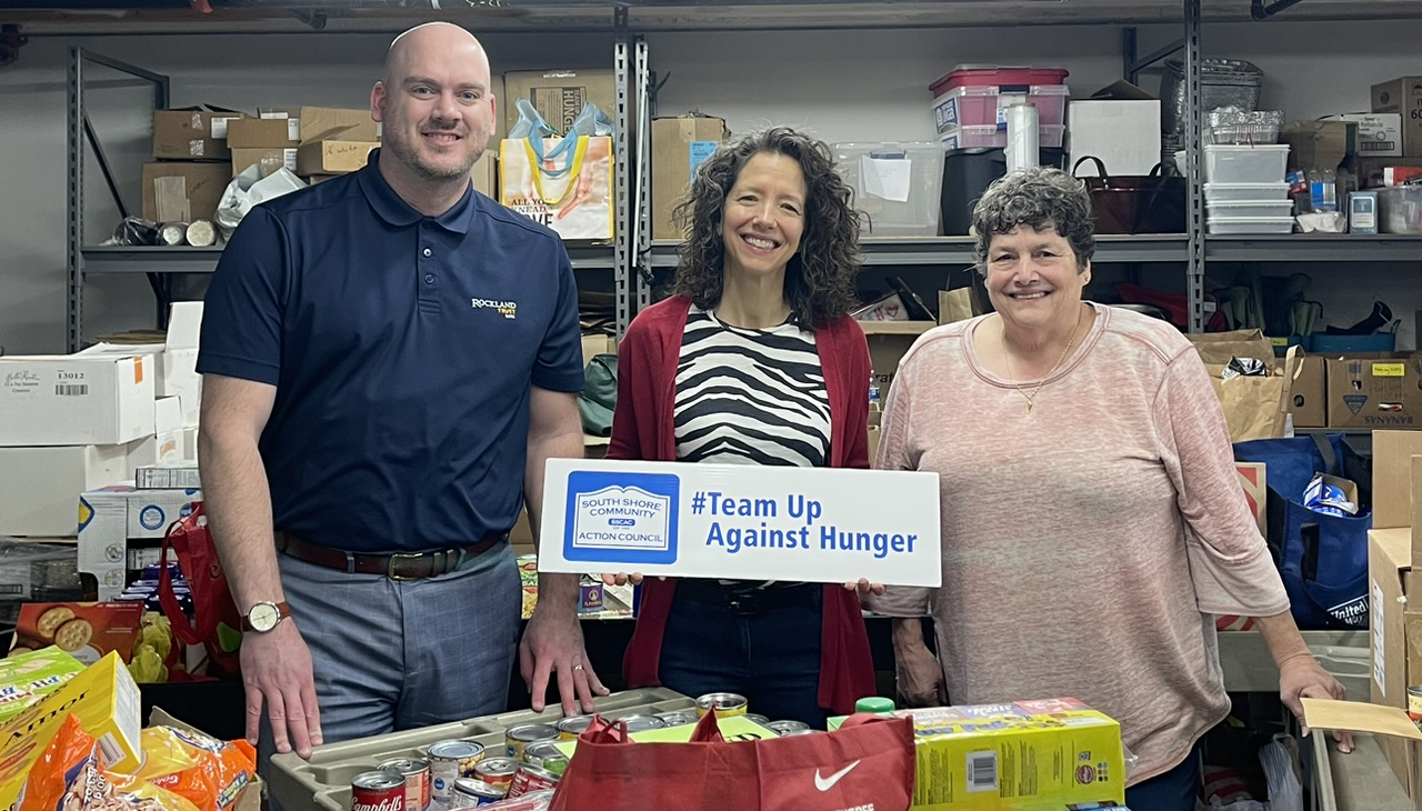 Three people stand in a storage room filled with food, holding a