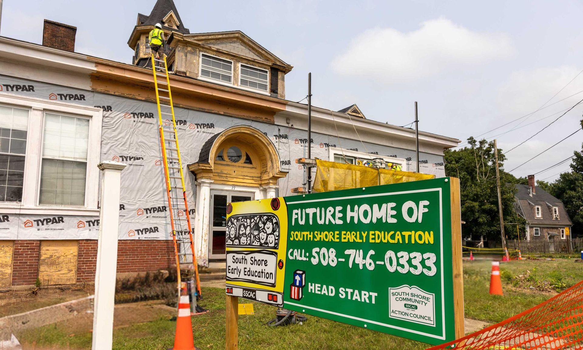A construction worker on a ladder works on the facade of a building behind a sign for North Shore Early Education.