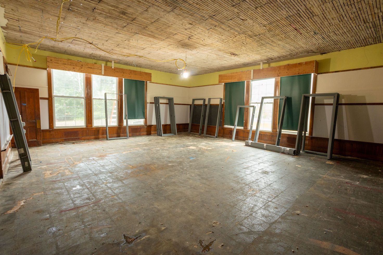 An empty, rustic room with a textured ceiling, worn wooden floors, tall windows, and several metal door frames leaning up.