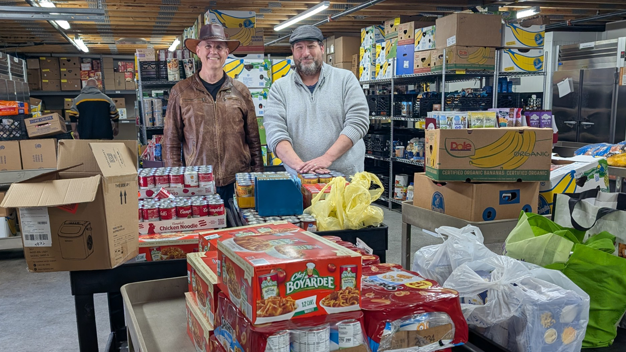 Two people stand in a food pantry warehouse surrounded by shelves of organized groceries, canned goods, and cardboard boxes.