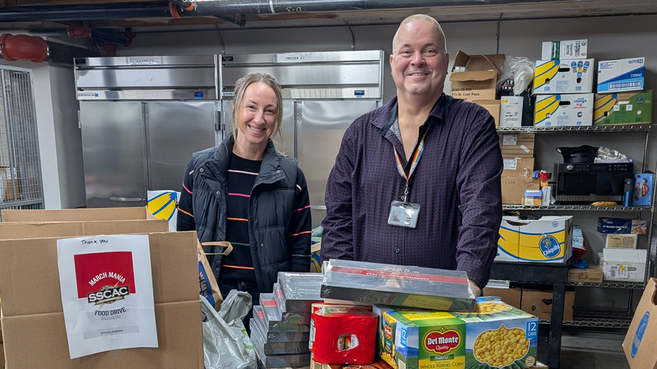 Two people stand behind a table with boxes of food in a storage area with metal shelving and refrigerators.