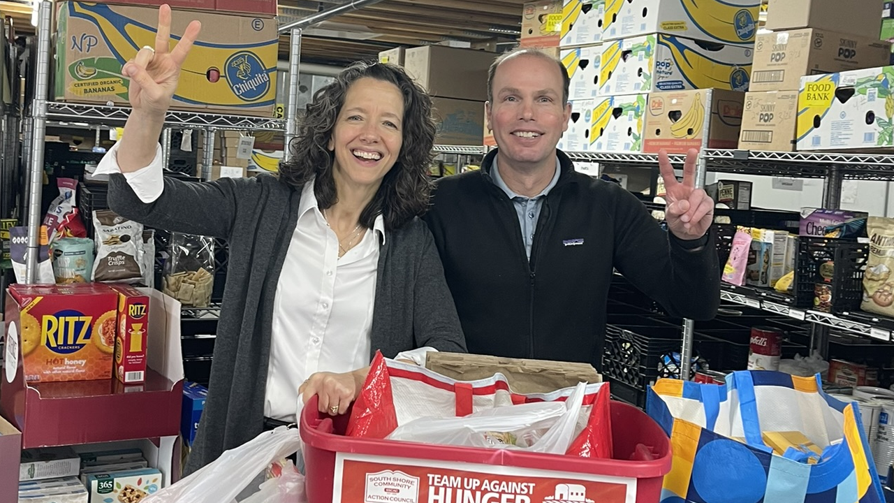 A smiling man and woman pose behind a food bank donation bin, with the woman making a peace sign in a warehouse setting.