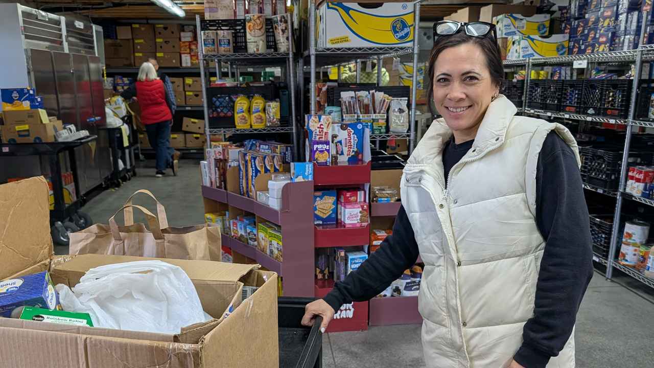 A person in a cream-colored puffer vest stands in a food pantry, holding a shopping cart near boxes of groceries.