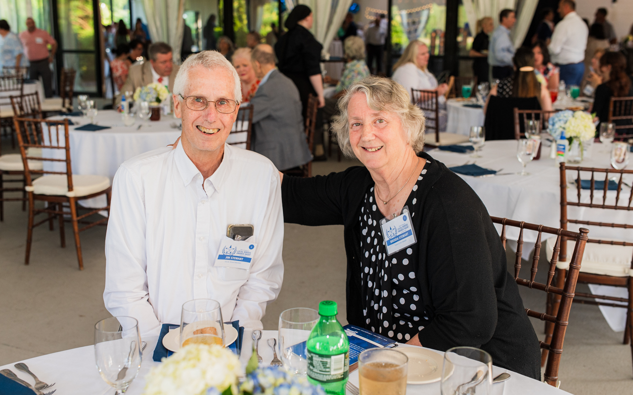 Two smiling people seated at a table in a bright banquet hall, with drinks and table settings nearby