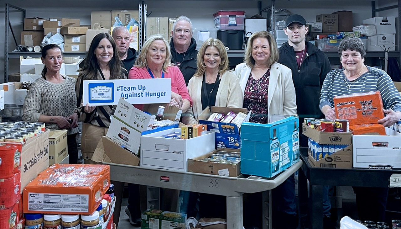 A group of people posing behind tables filled with boxes of food donations in a storage facility.
