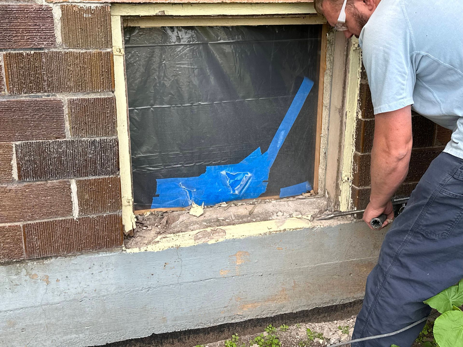 A person in a blue shirt works on an exterior window frame, removing material with a tool.