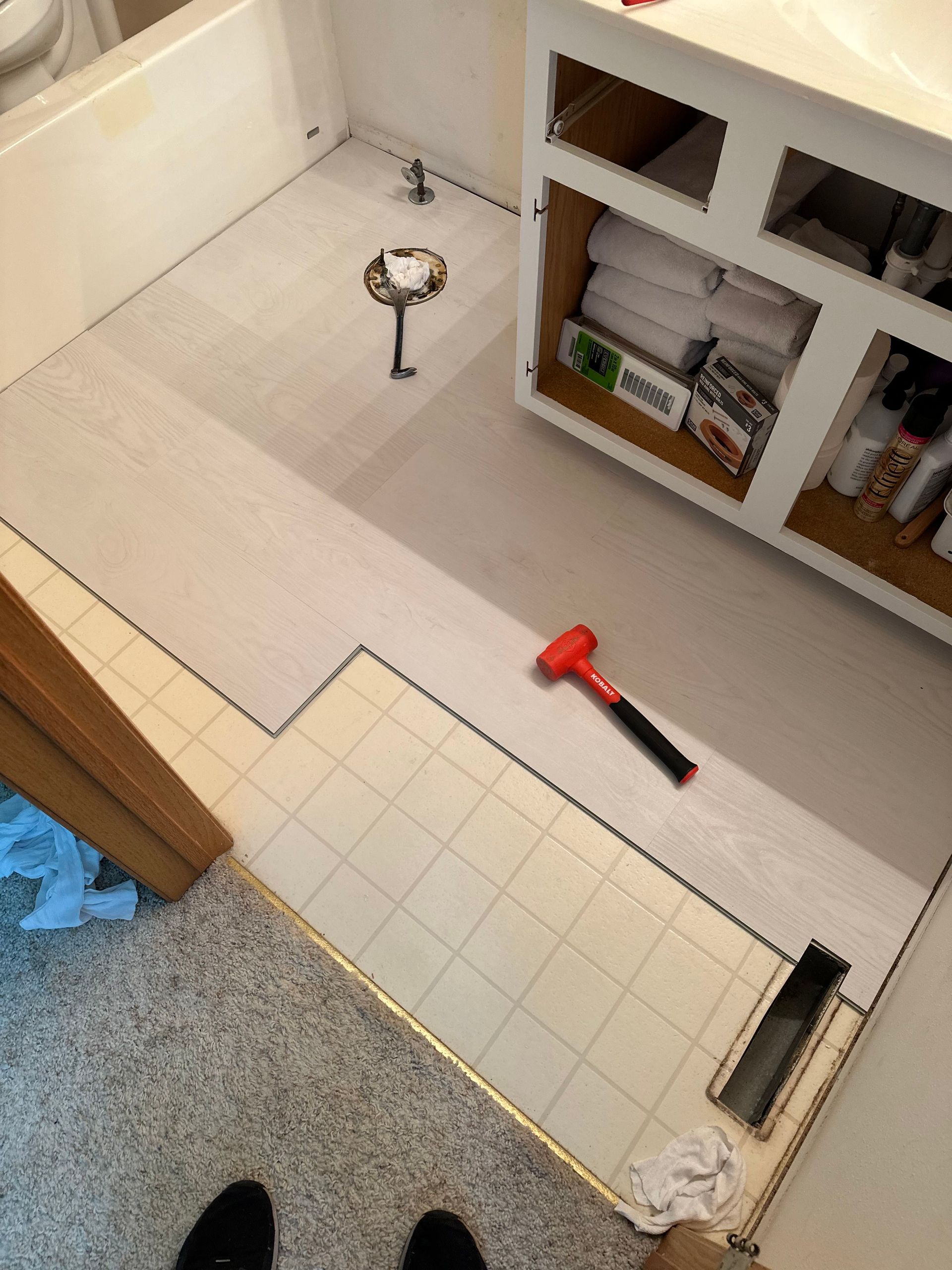 Bathroom floor renovation in progress. Light gray vinyl plank flooring installed next to existing cream-colored tile. 