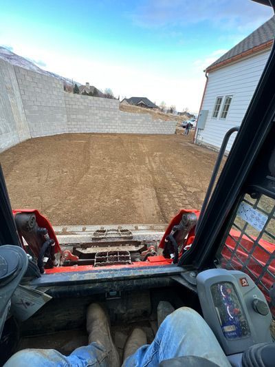 View from a skid steer cab over a dirt yard beside a white house and retaining wall under a blue sky