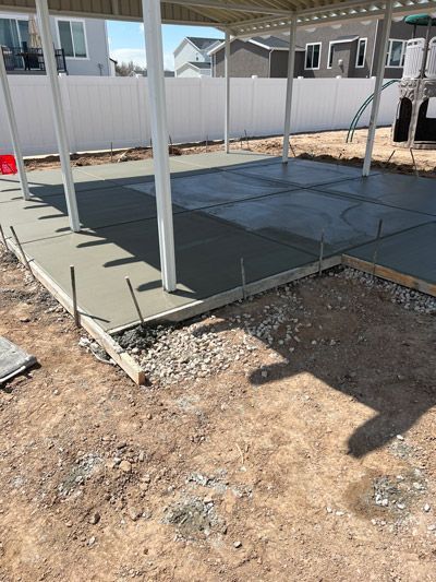 Concrete slab under construction beside a white house and fence, with a metal roof frame overhead