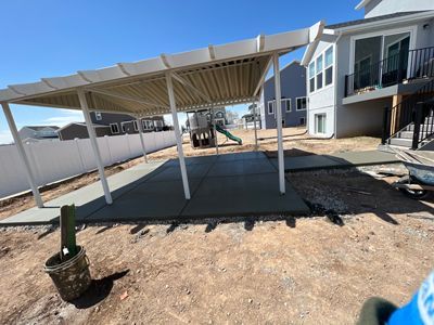 Covered carport in a dirt yard beside a modern house, with white posts and a potted plant in front.