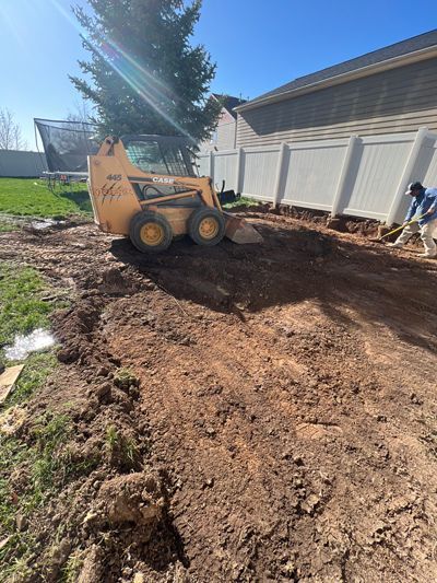 Construction skid steer leveling dirt beside a white garage in a sunny backyard