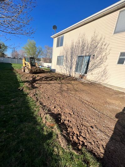 Excavator grading soil beside a beige house under a clear blue sky
